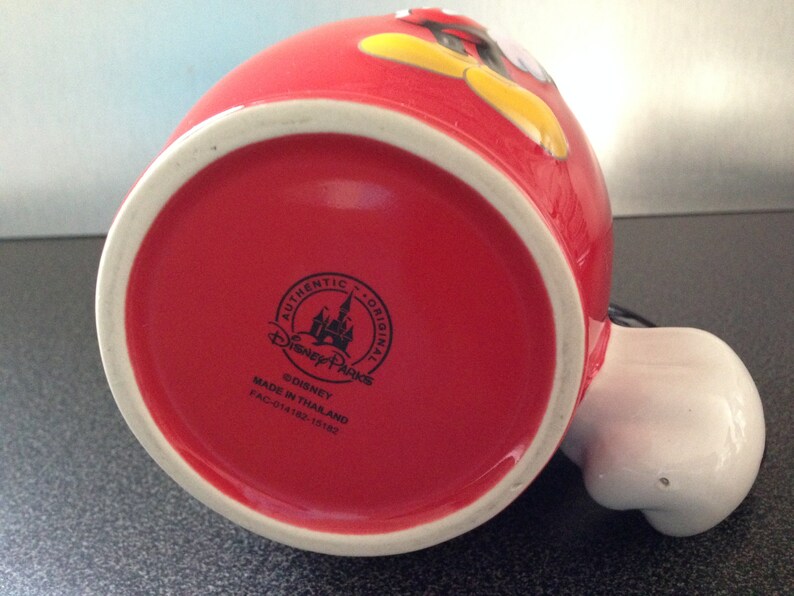 May include: Red ceramic mug with a cartoon image of Mickey Mouse. The mug has a white rim and a black Disney Parks logo on the bottom. The logo reads "Authentic Original Disney Parks &copy;Disney Made in Thailand FAC-014182-15182."