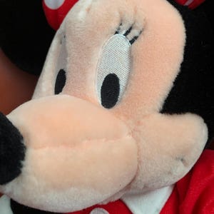 Może przedstawiać: Zbliżenie pluszowej zabawki Minnie Mouse w czerwonej sukience w białe kropki. Jej twarz jest r&oacute;żowa, a oczy i rzęsy czarne.