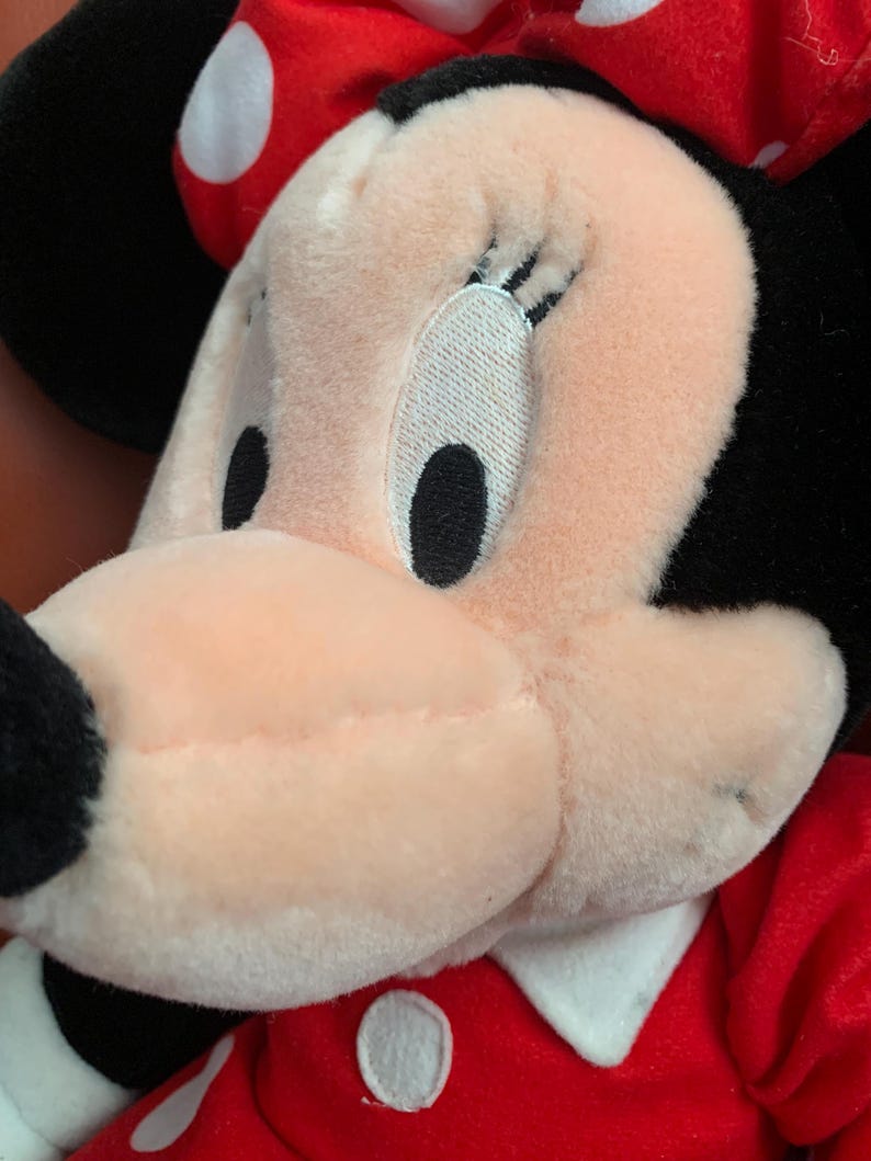 Może przedstawiać: Zbliżenie pluszowej zabawki Minnie Mouse w czerwonej sukience w białe kropki. Jej twarz jest r&oacute;żowa, a oczy i rzęsy czarne.