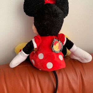 May include: A plush Minnie Mouse toy wearing a red dress with white polka dots and a red bow. The toy has a black tail and is sitting on a brown sofa. The tag on the toy reads "Authentic Original Disney Parks".