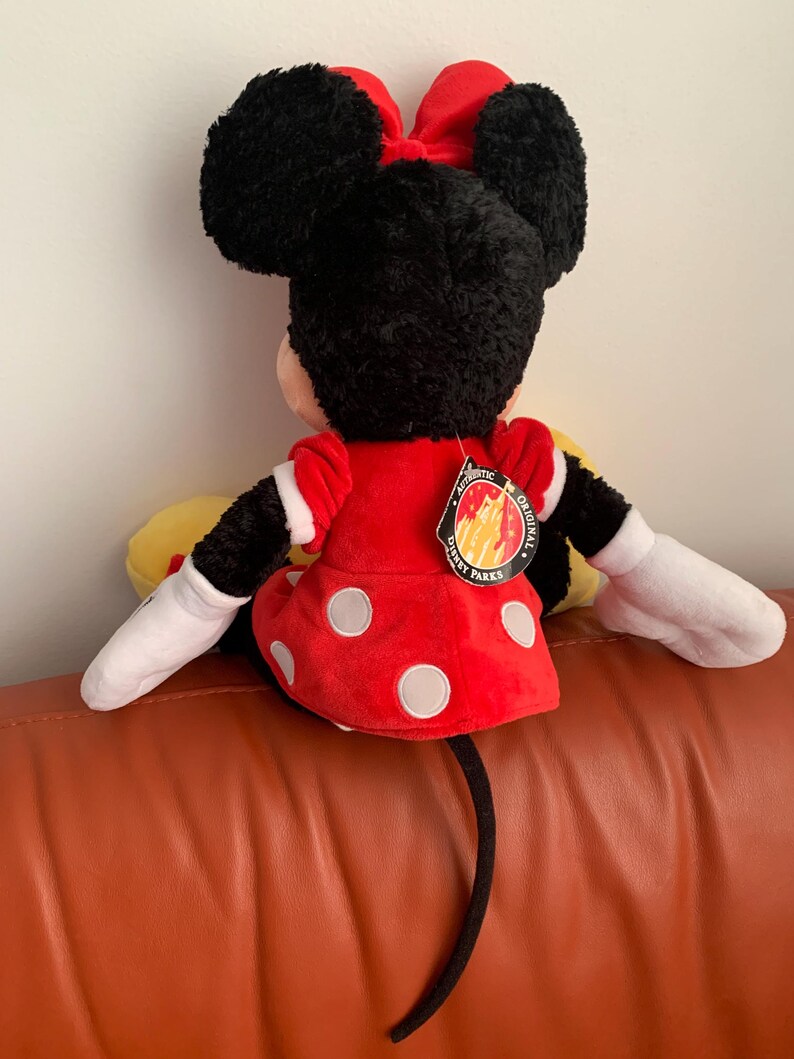May include: A plush Minnie Mouse toy wearing a red dress with white polka dots and a red bow. The toy has a black tail and is sitting on a brown sofa. The tag on the toy reads "Authentic Original Disney Parks".