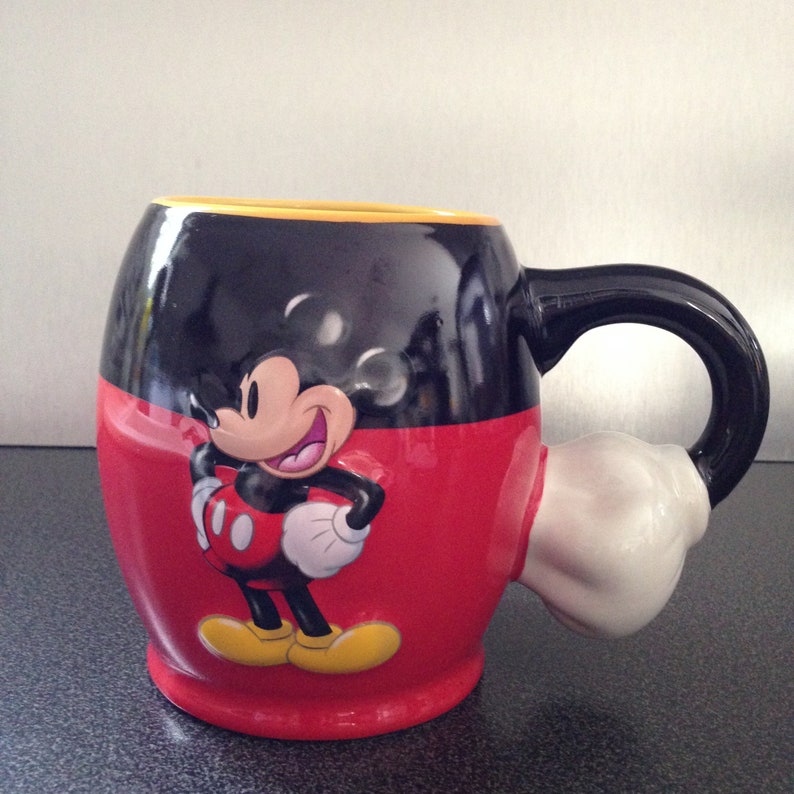 May include: A red and black ceramic mug with a Mickey Mouse character design. The mug has a handle shaped like Mickey Mouse's hand.