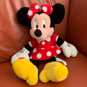 Może przedstawiać: Pluszowa zabawka Minnie Mouse w czerwonej sukience w białe kropki i czerwoną kokardkę w białe kropki. Zabawka ma ż&oacute;łte stopy i siedzi na brązowej kanapie.
