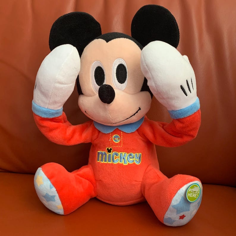 Puede incluir: Un juguete de peluche Mickey Mouse rojo y azul que lleva un pijama con la palabra "Mickey" en la parte delantera. El juguete tiene un bot&oacute;n en la parte inferior que dice "&iexcl;Presiona aqu&iacute;!"