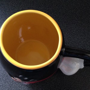 May include: Black ceramic mug with a yellow rim and a white handle. The mug features a cartoon character with a red nose and black eyes.