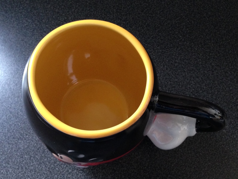 May include: Black ceramic mug with a yellow rim and a white handle. The mug features a cartoon character with a red nose and black eyes.