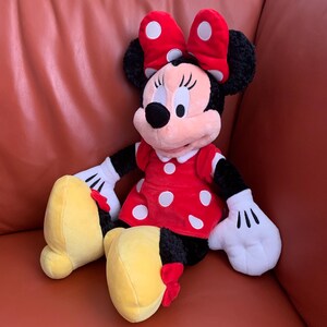 May include: A large plush Minnie Mouse toy wearing a red dress with white polka dots and a red bow with white polka dots. The toy has yellow shoes and is sitting on a brown leather sofa.