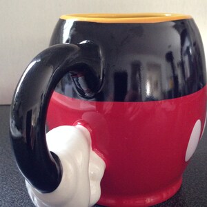 May include: A black and red ceramic mug with a white glove-shaped handle. The mug is decorated with a black outline of Mickey Mouse's head and two white circles.