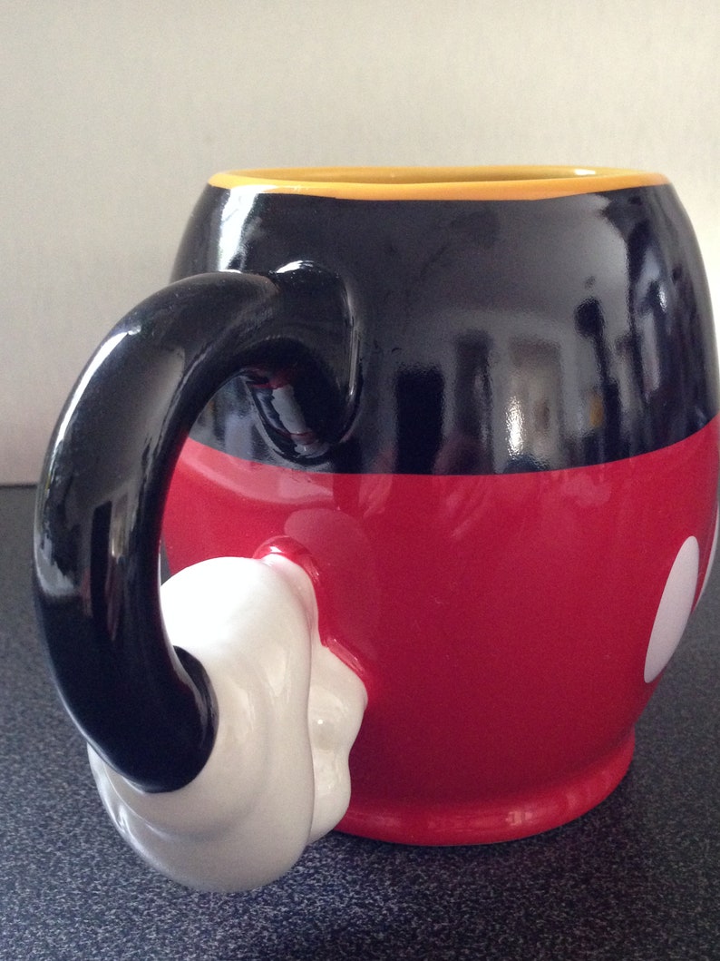 May include: A black and red ceramic mug with a white glove-shaped handle. The mug is decorated with a black outline of Mickey Mouse's head and two white circles.