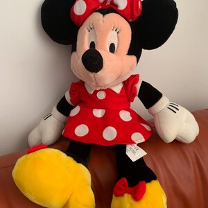 Może przedstawiać: Pluszowa zabawka przedstawiająca Minnie Mouse w czerwonej sukience w białe kropki i z czerwoną kokardką w białe kropki. Ma czarne uszy i czarny nos.