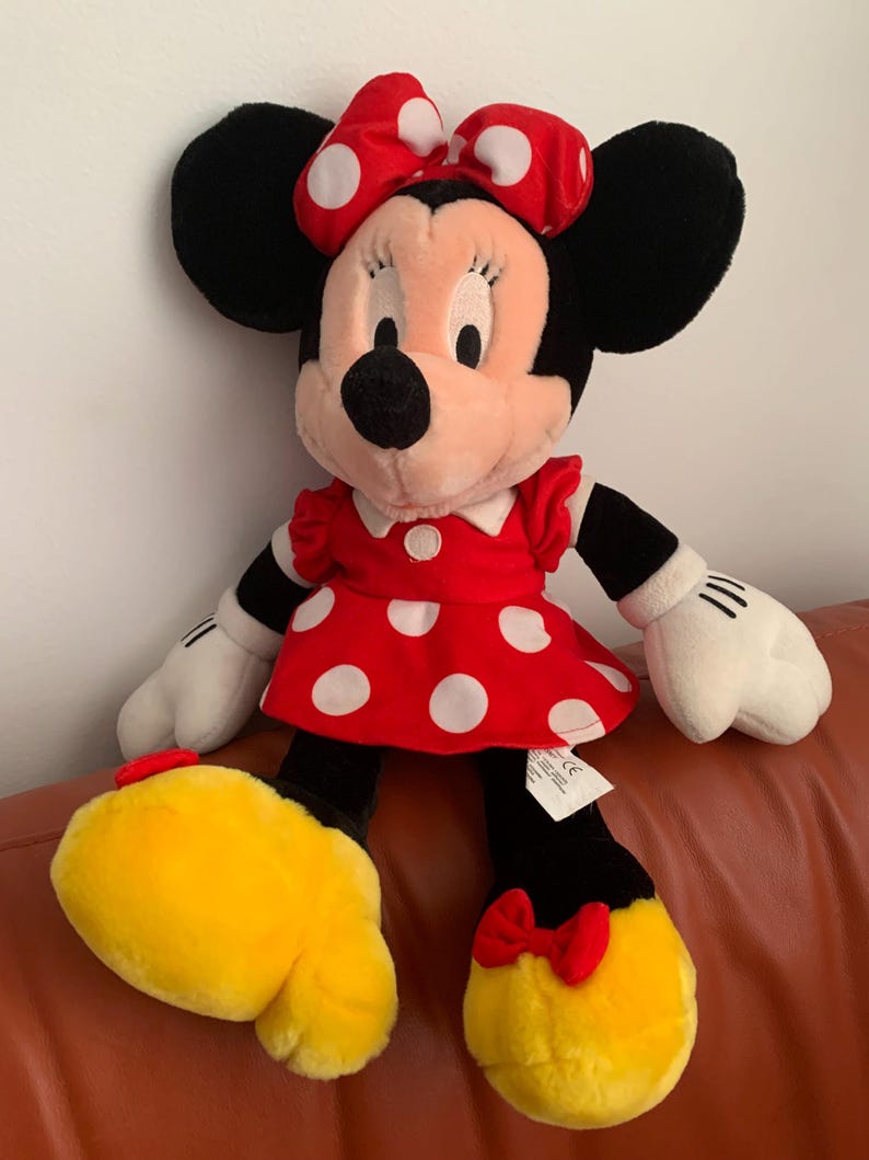 Może przedstawiać: Pluszowa zabawka przedstawiająca Minnie Mouse w czerwonej sukience w białe kropki i z czerwoną kokardką w białe kropki. Ma czarne uszy i czarny nos.