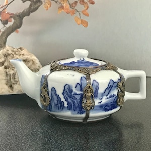 May include: A white porcelain teapot with a blue and white landscape design. The teapot is decorated with a metal band and has a lid with a knob.