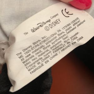 Może przedstawiać: Biała metka z czarnym tekstem, na kt&oacute;rej widnieje napis "The Walt Disney Company &copy;DISNEY" oraz informacje o materiałach, pochodzeniu i producencie produktu. Metka jest przymocowana do czarno-białej pluszowej zabawki.