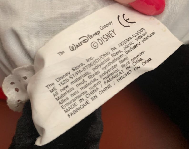 Może przedstawiać: Biała metka z czarnym tekstem, na kt&oacute;rej widnieje napis "The Walt Disney Company &copy;DISNEY" oraz informacje o materiałach, pochodzeniu i producencie produktu. Metka jest przymocowana do czarno-białej pluszowej zabawki.