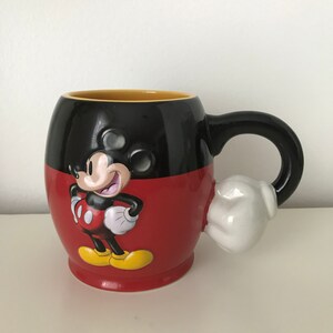 May include: A red and black ceramic mug with a 3D Mickey Mouse design. Mickey is wearing his signature red shorts and yellow shoes. The mug has a white handle shaped like Mickey's glove.