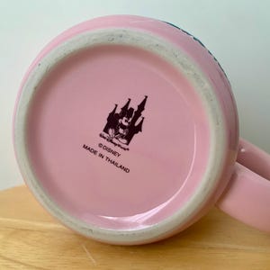 May include: A pink ceramic mug with a handle, viewed from the bottom. The mug features a black and white Disney logo with Mickey Mouse and a castle. The text "&copy;Disney MADE IN THAILAND" is printed below the logo.