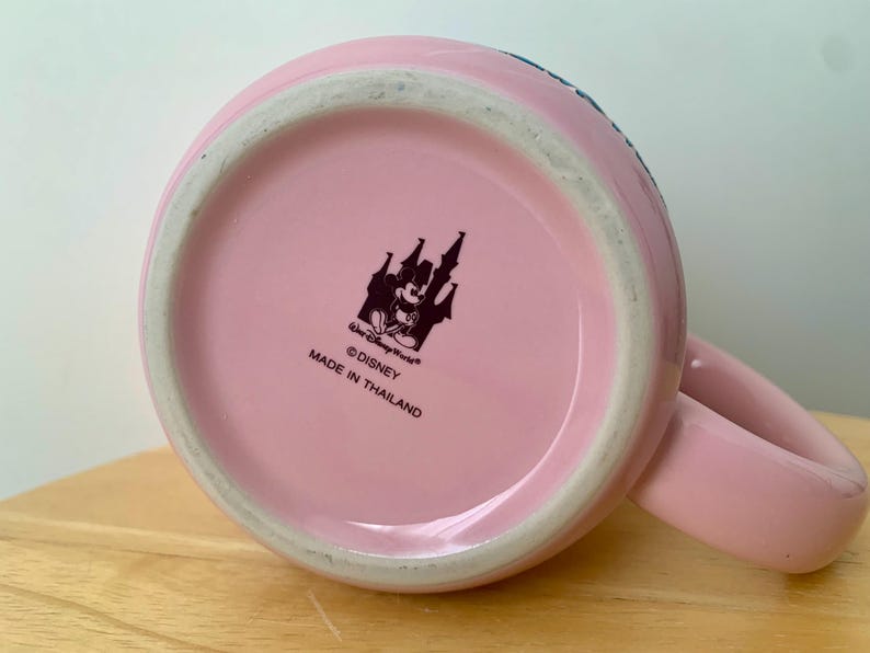 May include: A pink ceramic mug with a handle, viewed from the bottom. The mug features a black and white Disney logo with Mickey Mouse and a castle. The text "&copy;Disney MADE IN THAILAND" is printed below the logo.