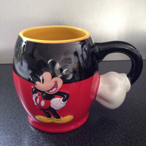 May include: A black and red ceramic mug with a Mickey Mouse design. The mug has a white handle shaped like Mickey Mouse's glove.