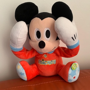 Puede incluir: Juguete de peluche Mickey Mouse rojo con pijama de ribete azul y blanco. El juguete tiene un bot&oacute;n en el pie que dice "Presiona aqu&iacute;".