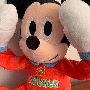Puede incluir: Un juguete de peluche de Mickey Mouse con un atuendo rojo y azul. El juguete tiene un gran "mickey" bordado en la parte delantera de su pecho.