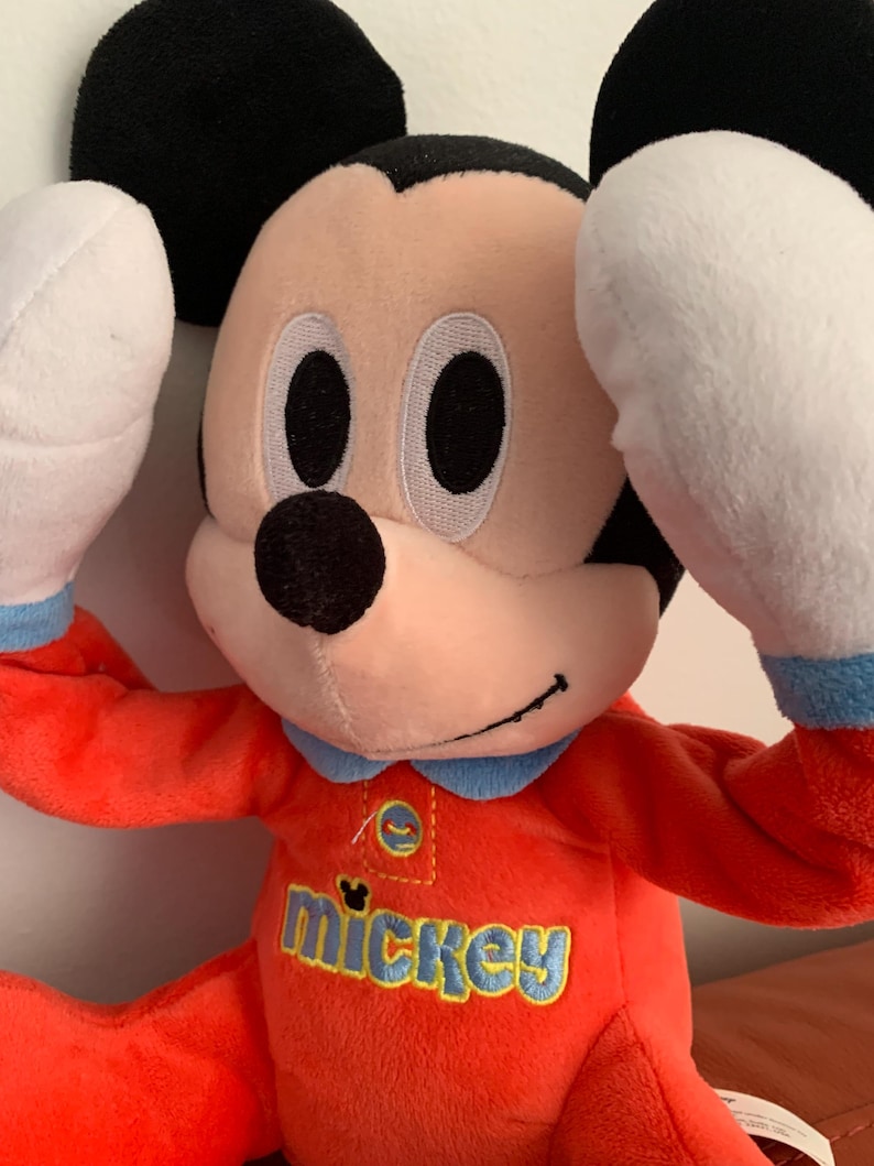 Puede incluir: Un juguete de peluche de Mickey Mouse con un atuendo rojo y azul. El juguete tiene un gran "mickey" bordado en la parte delantera de su pecho.