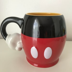 May include: A red and black ceramic mug with a white handle shaped like Mickey Mouse's glove. The mug has two white circles on the front, resembling Mickey Mouse's eyes.