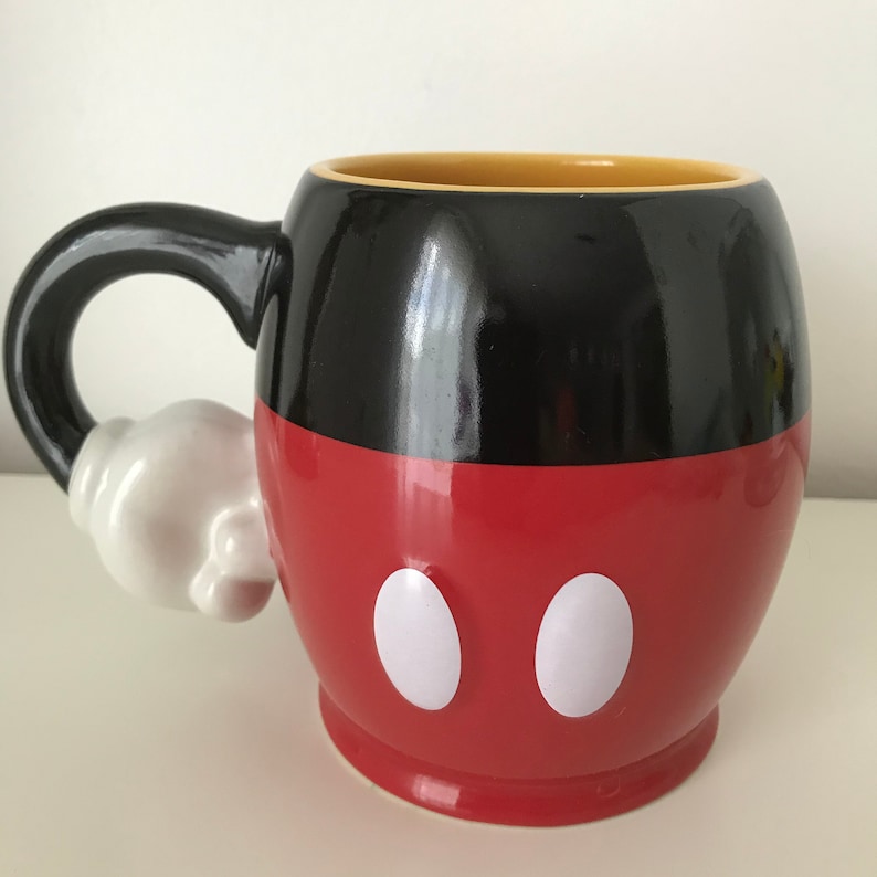 May include: A red and black ceramic mug with a white handle shaped like Mickey Mouse's glove. The mug has two white circles on the front, resembling Mickey Mouse's eyes.