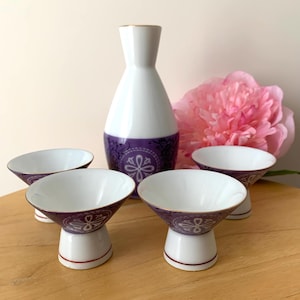 Peut inclure: Un service à saké blanc et violet comprenant une bouteille et quatre tasses assorties. La bouteille a un bord doré et une bande violette avec un motif floral. Les tasses ont un design similaire et sont blanches à l'intérieur. Une fleur rose est en arrière-plan.