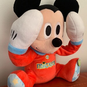 Puede incluir: Un juguete de peluche Mickey Mouse rojo que lleva un pijama azul y rojo con la palabra "mickey" en la parte delantera. El juguete tiene guantes blancos y est&aacute; sentado sobre una superficie roja.