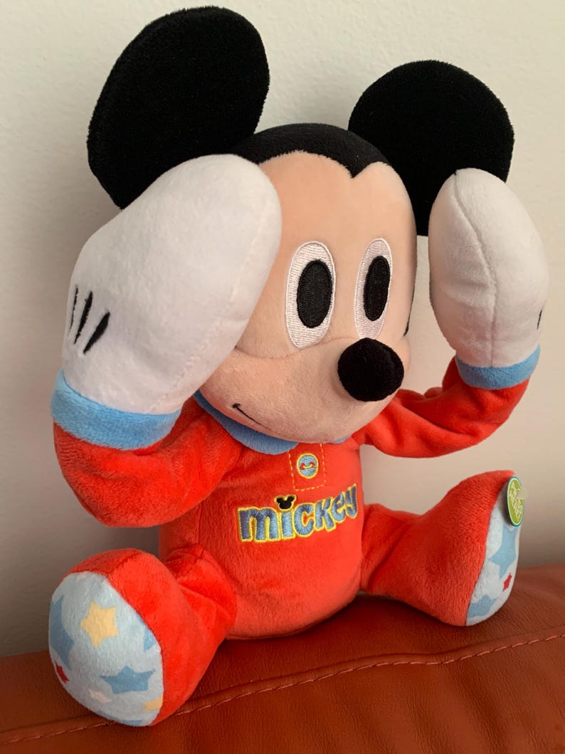 Puede incluir: Un juguete de peluche Mickey Mouse rojo que lleva un pijama azul y rojo con la palabra "mickey" en la parte delantera. El juguete tiene guantes blancos y est&aacute; sentado sobre una superficie roja.