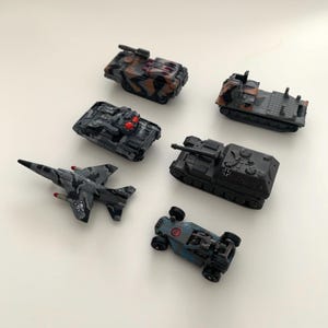 Puede incluir: Colección de vehículos militares de juguete, incluyendo tanques, un avión de combate y un camión. Los juguetes están pintados con patrones de camuflaje gris, negro y marrón. Un tanque tiene detalles en rojo. El avión tiene un diseño de calavera.