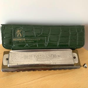 May include: A silver and brass harmonica with the text "SUPER CHROMONICA" and "CHROMATIC M. HOHNER HARMONICA". It is accompanied by a green, textured case with the text "HOHNER - Chromonica 270 MADE IN GERMANY".