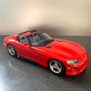 Puede incluir: Un coche modelo Dodge Viper rojo con interior negro. El coche de juguete tiene ruedas plateadas y un exterior detallado, que incluye faros, rejillas de ventilación y el logotipo de Viper. El coche es una réplica en miniatura de un coche deportivo.