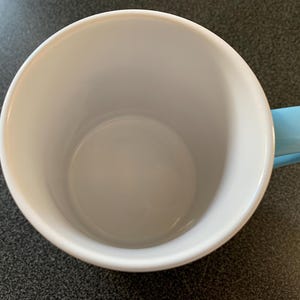 May include: A white ceramic mug with a light blue handle. The mug is empty and has a smooth, glossy finish.