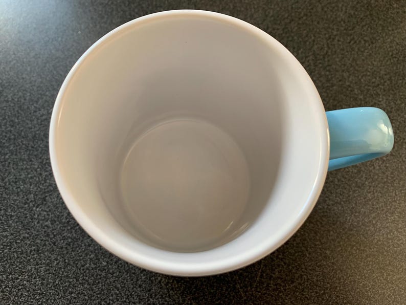 May include: A white ceramic mug with a light blue handle. The mug is empty and has a smooth, glossy finish.