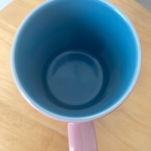 May include: A ceramic mug with a light blue interior and a pink handle. The rim is white, and the mug sits on a wooden surface. The mug is empty and viewed from above.
