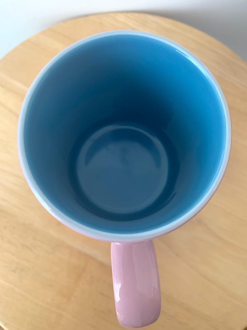 May include: A ceramic mug with a light blue interior and a pink handle. The rim is white, and the mug sits on a wooden surface. The mug is empty and viewed from above.