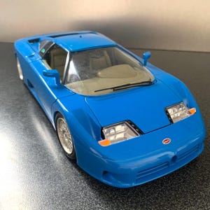 May include: A vibrant blue Bugatti model car with silver wheels. The toy car features a detailed interior and headlights. The Bugatti logo is displayed on the front. The car is a miniature replica of a sports car.