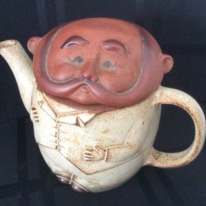 Może przedstawiać: Ceramiczny czajnik w kształcie człowieka z brązową głową i białym ciałem. Czajnik ma wąsy i białą koszulę z guzikami.