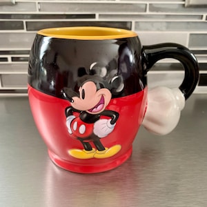 May include: A red and black ceramic mug with a 3D Mickey Mouse design. The mug has a yellow rim and a white handle shaped like Mickey Mouse's glove.