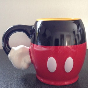 May include: A black and red ceramic mug with a white handle shaped like Mickey Mouse's glove. The mug has a yellow rim and two white circles on the front.