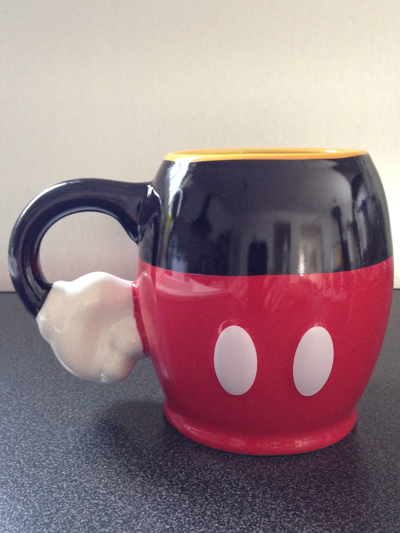 May include: A black and red ceramic mug with a white handle shaped like Mickey Mouse's glove. The mug has a yellow rim and two white circles on the front.