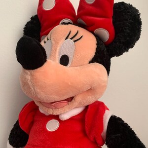May include: A plush red and black Minnie Mouse toy with a white polka dot bow and dress. The toy has a friendly expression and is made of soft, cuddly material.