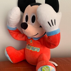 Puede incluir: Juguete de peluche Mickey Mouse rojo que lleva un pijama rojo y azul con la palabra "Mickey" en la parte delantera. El juguete tiene un guante blanco en una mano y un guante azul y blanco en la otra. El juguete tiene un pie azul y blanco con una estrella verde y las palabras "&iexcl;Presiona aqu&iacute;!" en &eacute;l.