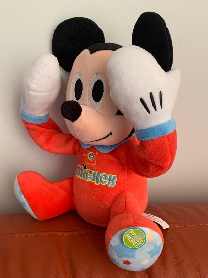 Puede incluir: Juguete de peluche Mickey Mouse rojo que lleva un pijama rojo y azul con la palabra "Mickey" en la parte delantera. El juguete tiene un guante blanco en una mano y un guante azul y blanco en la otra. El juguete tiene un pie azul y blanco con una estrella verde y las palabras "&iexcl;Presiona aqu&iacute;!" en &eacute;l.