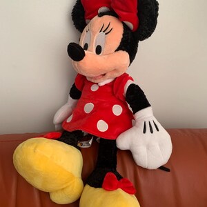 May include: A large plush toy of Minnie Mouse wearing a red dress with white polka dots and a red bow with white polka dots. She has black fur, a pink nose, and large black eyes.