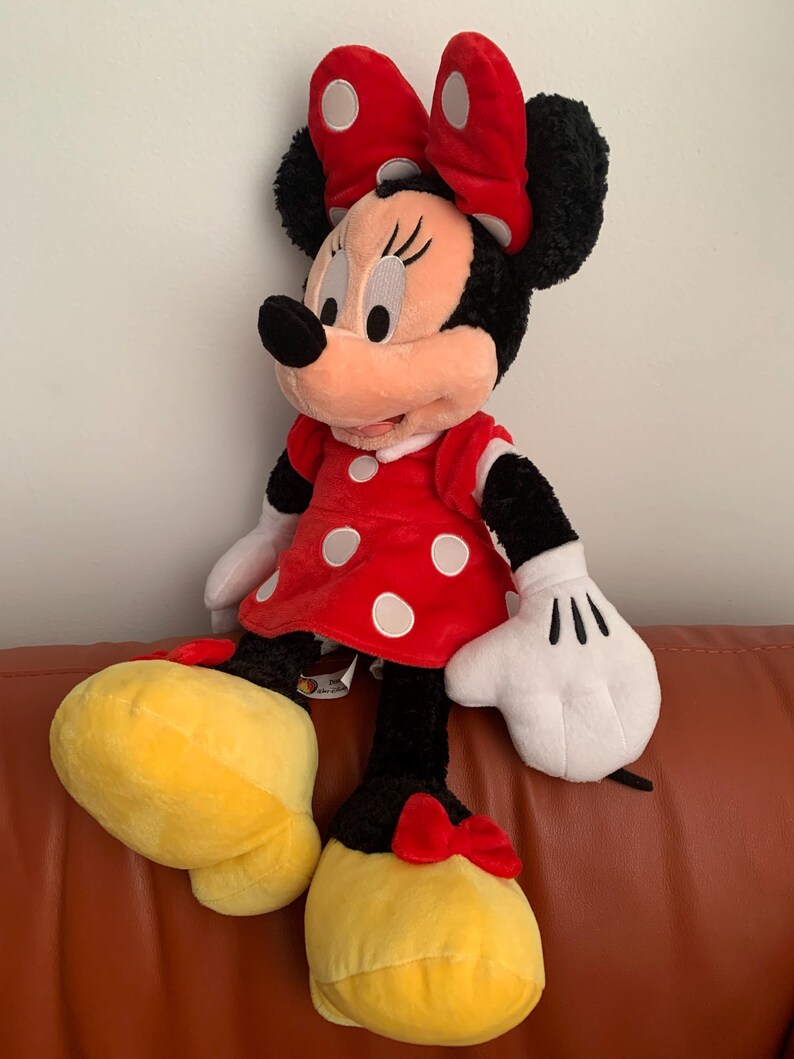May include: A large plush toy of Minnie Mouse wearing a red dress with white polka dots and a red bow with white polka dots. She has black fur, a pink nose, and large black eyes.