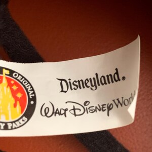 May include: White tag with black text that reads "Disneyland. Walt Disney World" and a yellow and red logo with a castle and stars that reads "Original Disney Parks".
