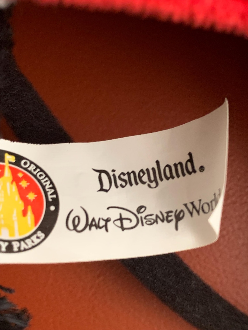 May include: White tag with black text that reads "Disneyland. Walt Disney World" and a yellow and red logo with a castle and stars that reads "Original Disney Parks".
