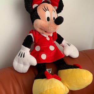 May include: A large plush toy of Minnie Mouse wearing a red dress with white polka dots and a red bow with white polka dots. She has black fur, a pink nose, and yellow shoes.